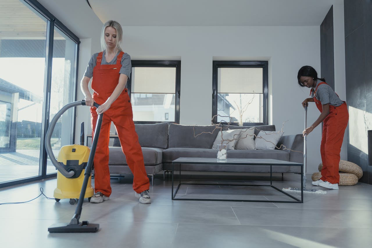 Two women in red overalls vacuuming and sweeping a modern living room.